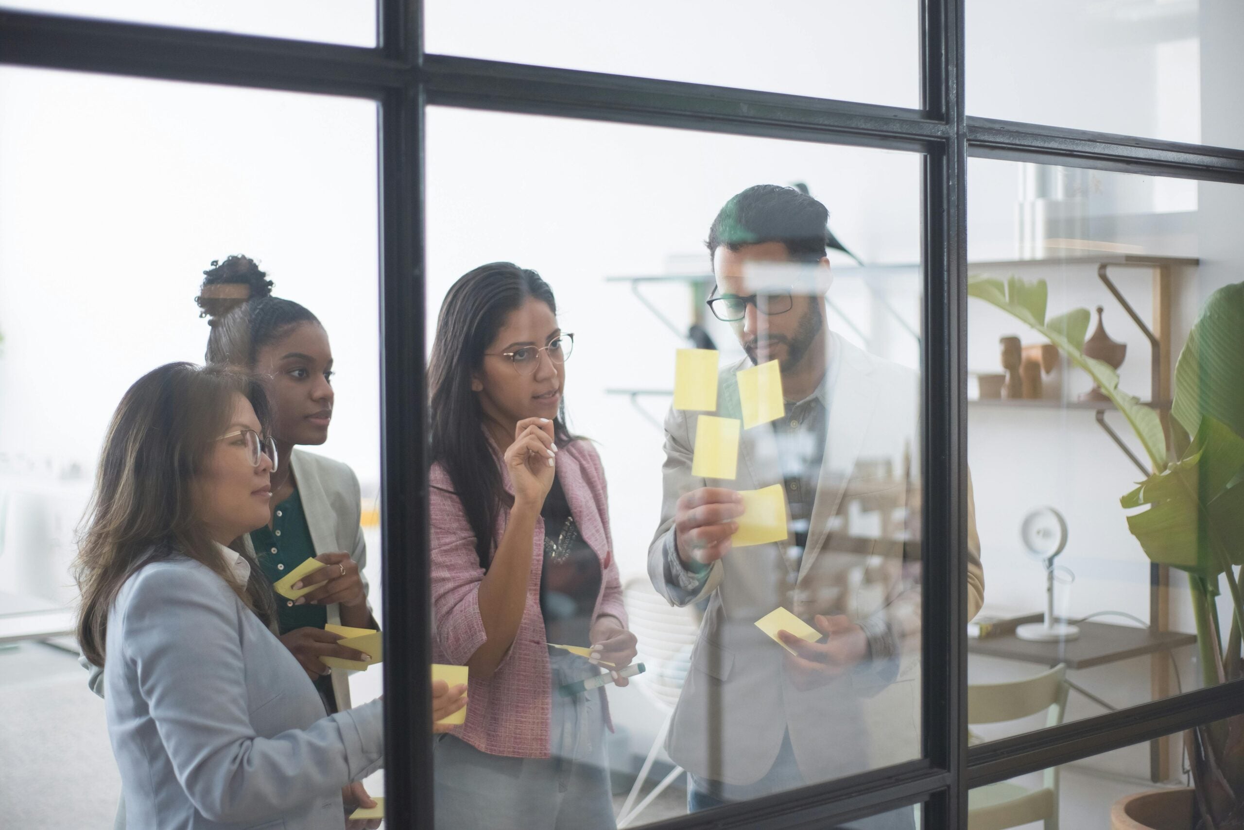 diverse group of colleagues in an office space look at a glass writing surface with post-its and writing on it