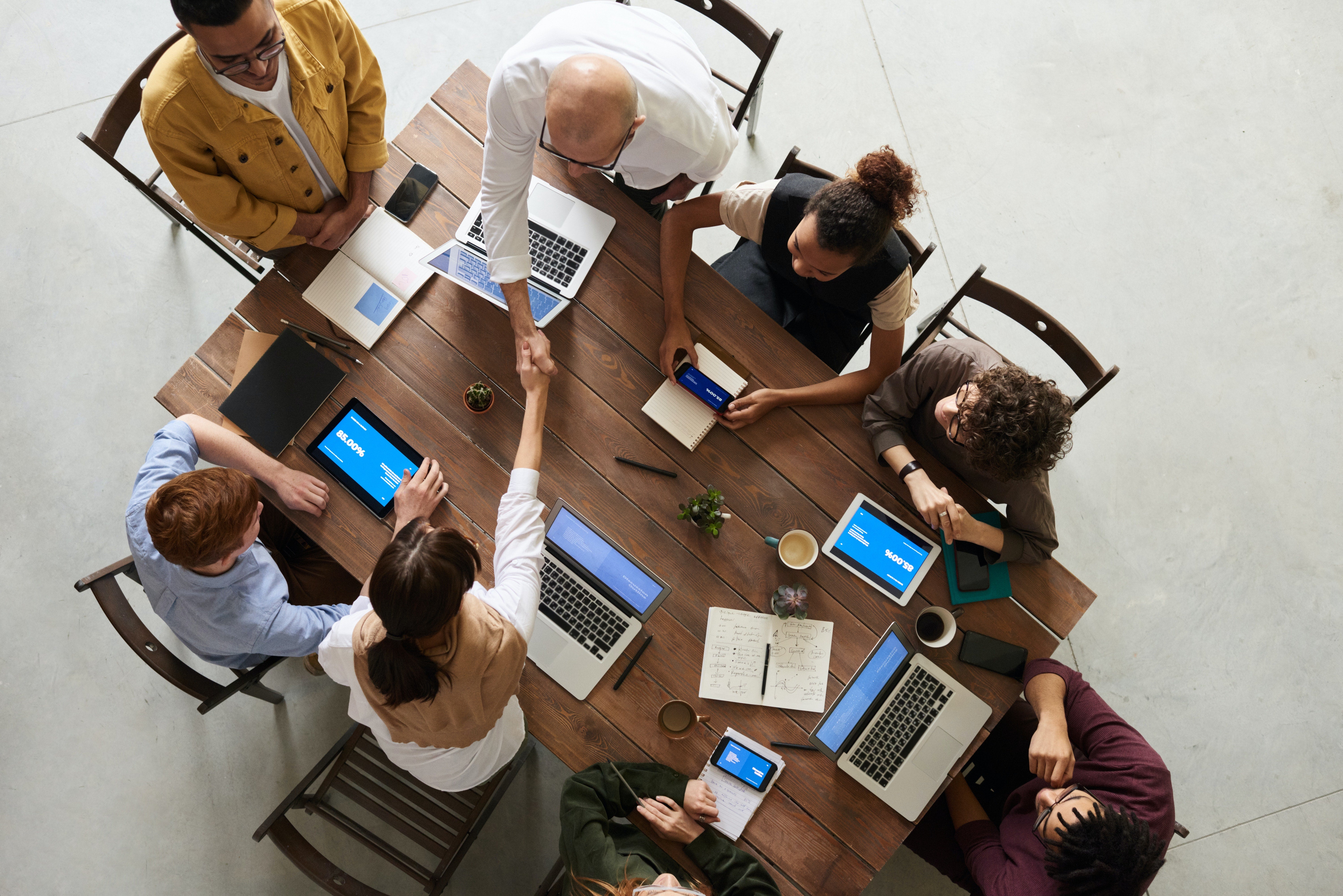 diverse group of colleagues in an office space look at a glass writing surface with post-its and writing on it, working on a desk with laptops and writing pads